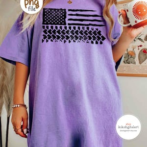May include: A light purple t-shirt featuring a distressed American flag design in black. The flag includes stars, stripes, and a tire track pattern. The shirt has short sleeves and a relaxed fit.