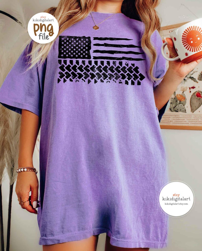 May include: A light purple t-shirt featuring a distressed American flag design in black. The flag includes stars, stripes, and a tire track pattern. The shirt has short sleeves and a relaxed fit.