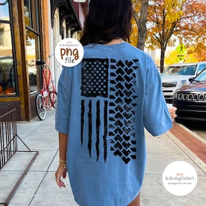 May include: A light blue t-shirt featuring a distressed American flag design. The flag's stars and stripes are depicted in black, with tire tracks replacing the stripes on the right side. The shirt has short sleeves and a relaxed fit.