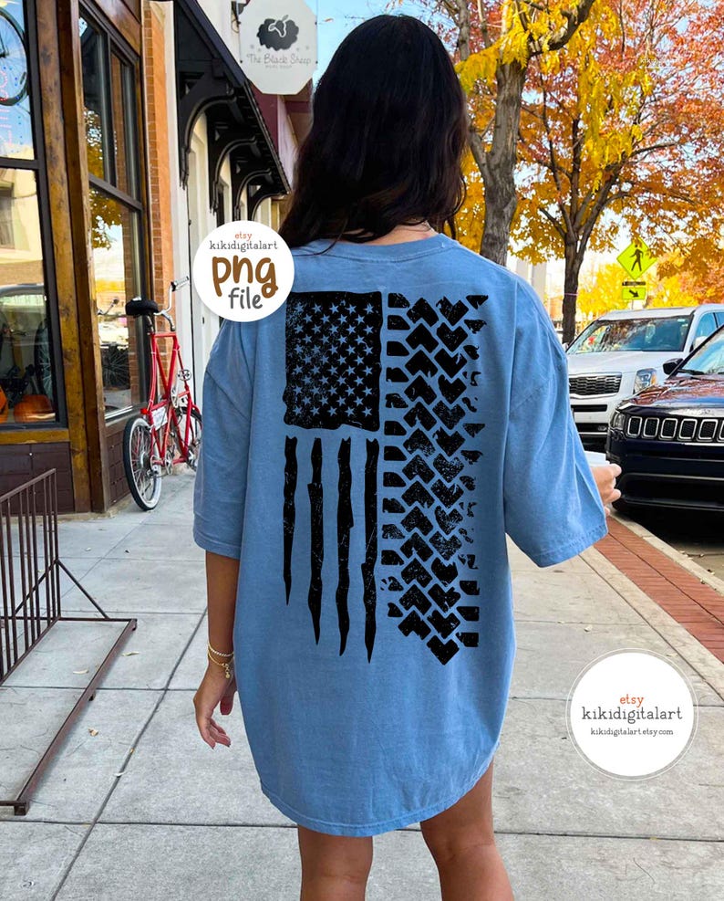 May include: A light blue t-shirt featuring a distressed American flag design. The flag's stars and stripes are depicted in black, with tire tracks replacing the stripes on the right side. The shirt has short sleeves and a relaxed fit.