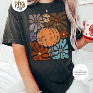 Op de afbeelding: Donkergrijs t-shirt met een pompoen- en bloemenontwerp in oranje, bruin, blauw en grijs. Het shirt heeft een vintage, distressed look. De Etsy shopnaam "kikidigitalart" is zichtbaar op het shirt.