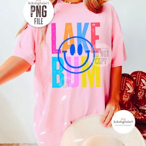 Op de afbeelding: Een roze T-shirt met een kleurrijk grafisch ontwerp met de tekst "Lake Bum" en een smiley in het midden. De grafiek heeft een verweerde look.