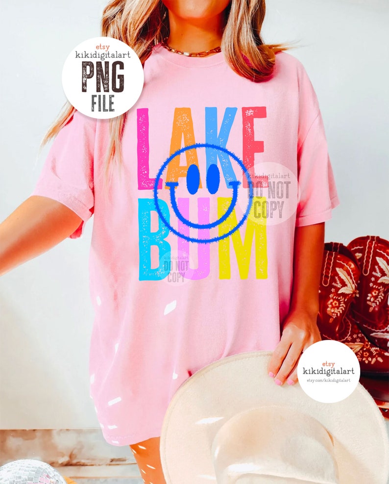Op de afbeelding: Een roze T-shirt met een kleurrijk grafisch ontwerp met de tekst "Lake Bum" en een smiley in het midden. De grafiek heeft een verweerde look.