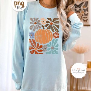 Op de afbeelding: Een lichtblauwe sweatshirt met een retro bloemen- en pompoenontwerp. De afbeelding bevat een oranje pompoen omringd door bloemen in bruin, oranje en blauw. De sweatshirt heeft lange mouwen en een ronde hals.