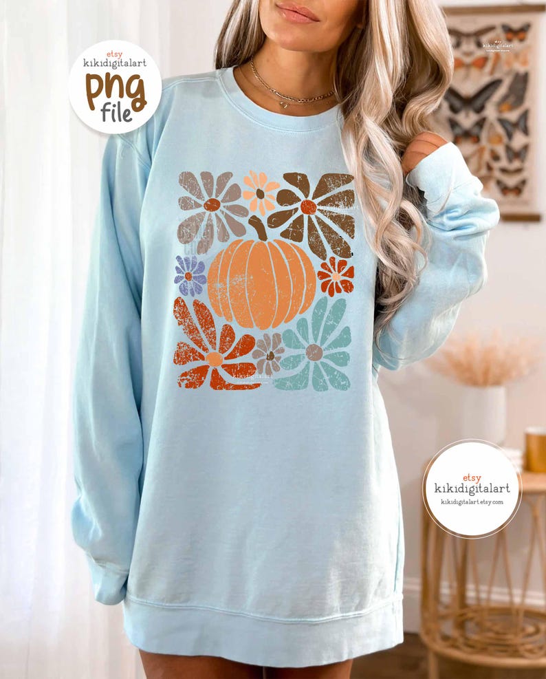 Op de afbeelding: Een lichtblauwe sweatshirt met een retro bloemen- en pompoenontwerp. De afbeelding bevat een oranje pompoen omringd door bloemen in bruin, oranje en blauw. De sweatshirt heeft lange mouwen en een ronde hals.