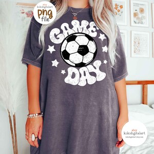 May include: A gray t-shirt with the words "GAME DAY" in white, surrounding a black and white soccer ball graphic. White stars are scattered around the text. The shirt has short sleeves and a relaxed fit.