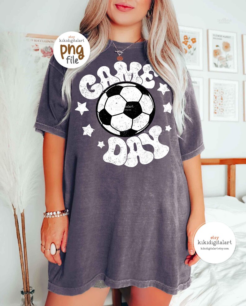 May include: A gray t-shirt with the words "GAME DAY" in white, surrounding a black and white soccer ball graphic. White stars are scattered around the text. The shirt has short sleeves and a relaxed fit.