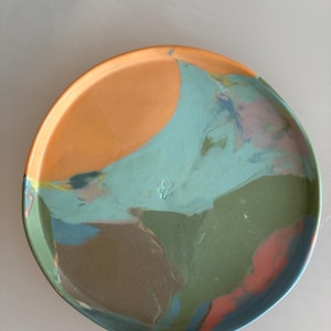 Puede incluir: Un plato redondo de cerámica con un patrón jaspeado en naranja, azul claro, verde oliva y rosa suave. La superficie tiene un acabado liso con un pequeño icono estampado en forma de flor situado cerca del centro de la base circular.