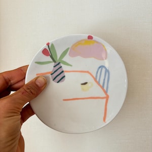 May include: A white ceramic plate with a whimsical illustration of a table setting. The design includes a pink and yellow dessert, a vase with flowers, a table, a chair, and a cup. The plate is held by a hand.