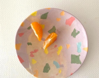 May include: A round, shallow bowl with a light lavender base featuring a multi-coloured terrazzo pattern of green, yellow, orange, pink, and light blue flecks. Two bright orange citrus segments rest inside the bowl on the interior surface.