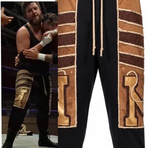 May include: Black sweatpants featuring tan suede-like side panels with horizontal dark brown textured stripes and thick cream-colored rope detailing. The pants include a matching cream drawstring at the waist and large gold-colored letter patches.