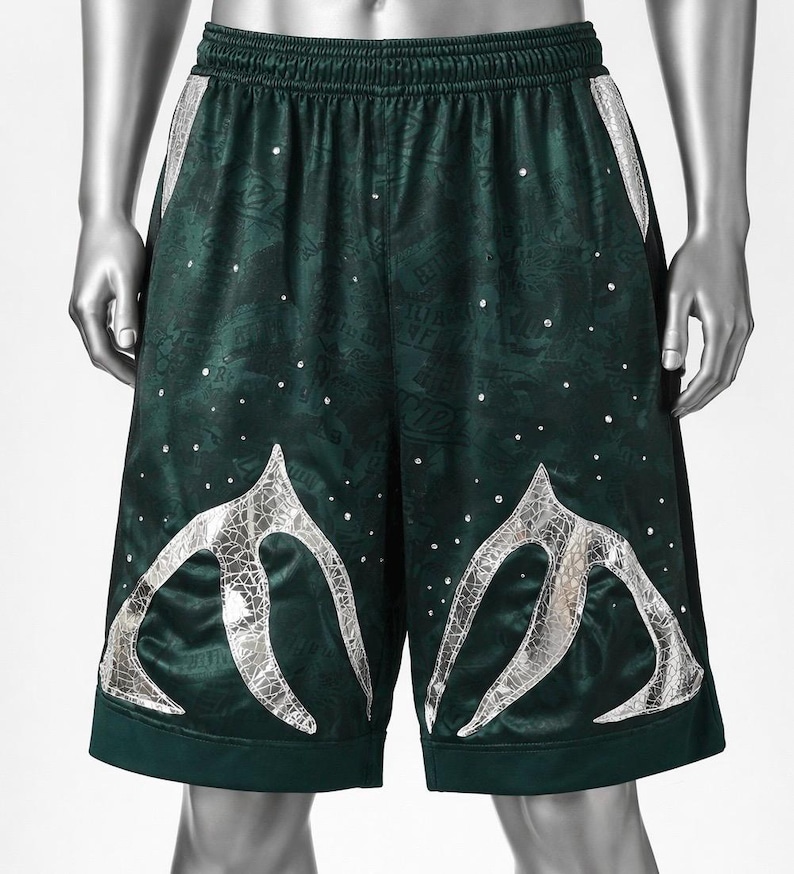 May include: Dark green athletic shorts featuring an elastic waistband, a subtle tonal pattern throughout the fabric, scattered small clear rhinestones, and two large, silver, cracked-texture graphic appliques on the lower front panels.