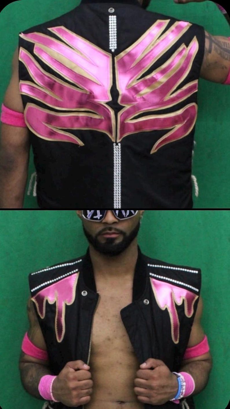 May include: A black sleeveless vest featuring metallic pink flame-like appliques on the chest and back. The garment includes vertical rows of silver rhinestones along the center back and shoulder panels, with a front zipper closure and silver studs.