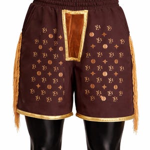 May include: Dark brown shorts featuring an elastic waistband, a central velvet panel, and gold-toned trim along the hem. The fabric is decorated with a repeating pattern of gold-colored symbols and letters, complemented by long gold fringe on the sides.