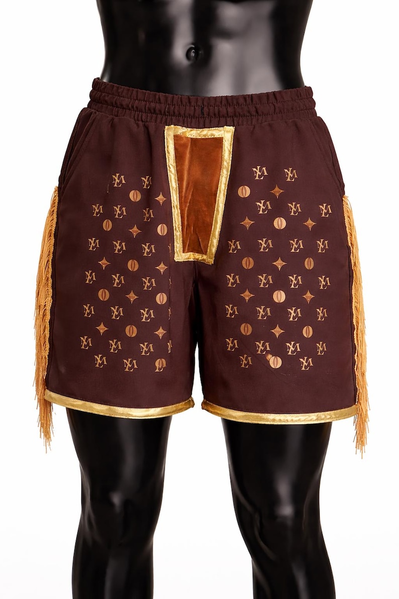 May include: Dark brown shorts featuring an elastic waistband, a central velvet panel, and gold-toned trim along the hem. The fabric is decorated with a repeating pattern of gold-colored symbols and letters, complemented by long gold fringe on the sides.