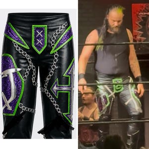 May include: Black faux leather wrestling pants featuring purple glitter panels, bright green trim, and silver metallic cross accents. The pants are decorated with silver chain details and a central front patch with two white X symbols.