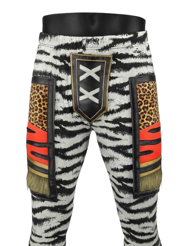 May include: White pants with a black zebra stripe pattern featuring a central black shield patch with two white X symbols. The sides include rectangular panels with leopard print, bright orange sections, gold trim, and olive green fringe accents.