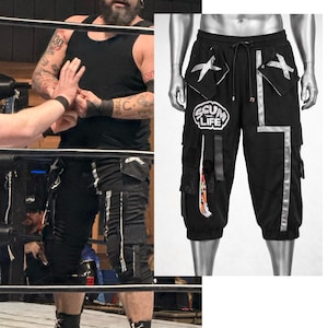 May include: Black cargo shorts featuring a drawstring waist, multiple pockets with silver X-shaped accents, and a prominent circular patch on the right leg displaying the text SCUM LIFE. The shorts include vertical silver reflective-style strips.