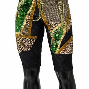 May include: Black shorts featuring a patchwork design of green sequins, leopard print fabric, and gold metallic mesh. The panels are separated by gold velvet trim and accented with hanging green fringe details throughout the front surface.