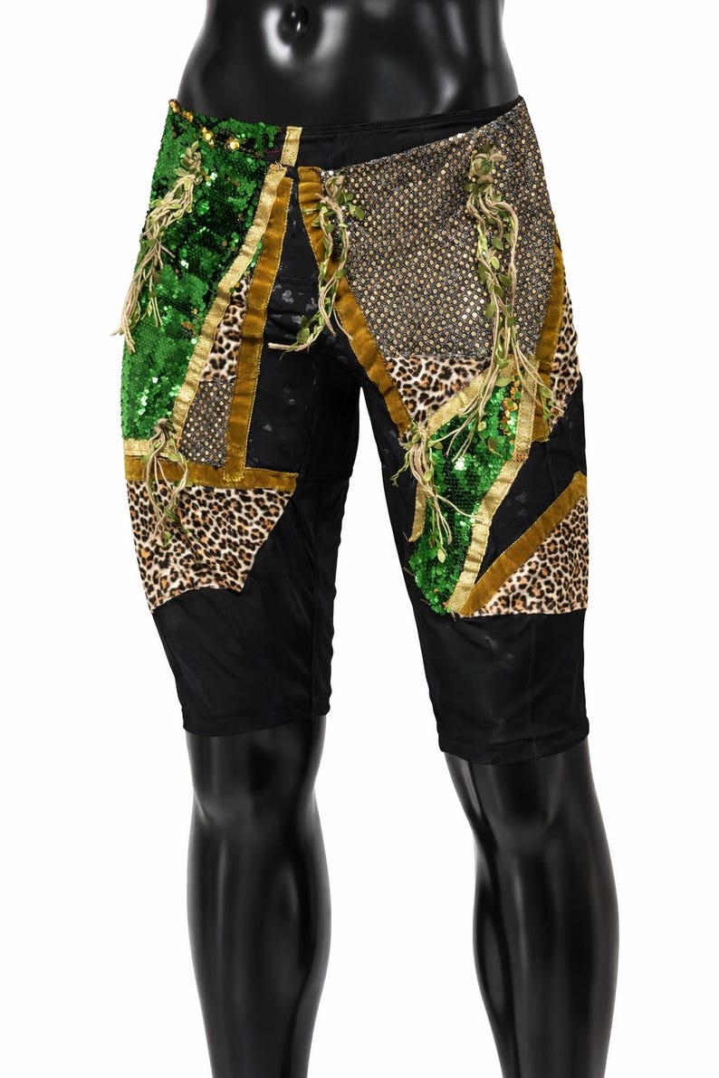 May include: Black shorts featuring a patchwork design of green sequins, leopard print fabric, and gold metallic mesh. The panels are separated by gold velvet trim and accented with hanging green fringe details throughout the front surface.