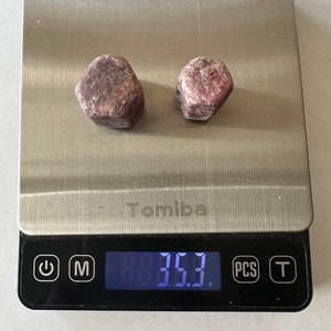 May include: Two pink and red rough gemstones on a digital scale that reads 3.53 grams. The scale is silver and black with the brand name "Tomiba" printed on it.