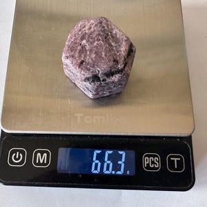 May include: A rough, hexagonal-shaped purple and grey mineral specimen resting on a digital kitchen scale. The scale features a stainless steel platform and a black digital display showing a weight measurement of 66.3 grams.