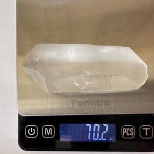 Puede incluir: Un cristal de cuarzo transparente que pesa 70,2 gramos en una báscula digital. La báscula tiene una pantalla negra con números blancos y una plataforma de metal plateado.