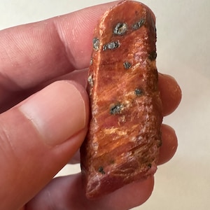 May include: A rough, red ruby crystal with black inclusions. The crystal is a deep red color and has a natural, unpolished finish.