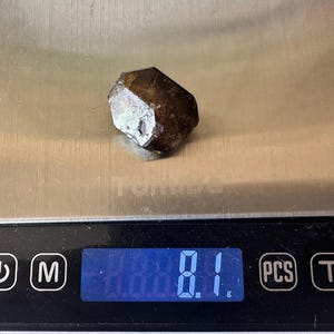 Op de afbeelding: Een donkerbruin, gefacetteerd kristal ligt op een digitale weegschaal. De weegschaal geeft een gewicht van 8,1 gram aan. Het kristal heeft een ruw, getextureerd oppervlak en lijkt een natuurlijk mineraal te zijn.
