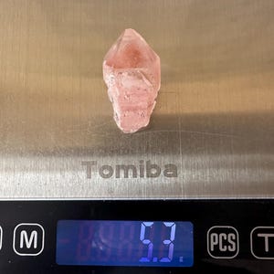 May include: A pink crystal sits on a digital scale, which reads 5.3 grams. The crystal is a pale pink hue and has a pointed end. The scale is silver and branded with "Tomiba".