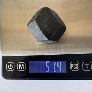 Op de afbeelding: Een donkere, facetgeslepen, zwarte edelsteen ligt op een digitale weegschaal en weegt 51,4 gram. De weegschaal heeft een zwarte basis met een digitale weergave van het gewicht. De edelsteen heeft een ruw, getextureerd oppervlak.