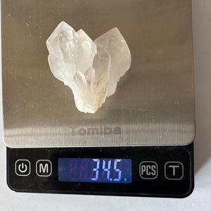 Puede incluir: Un grupo de cristales de cuarzo transparente descansa sobre una balanza digital, mostrando un peso de 34,5. La balanza es negra con una superficie de pesaje plateada. La marca "Tomiba" es visible.