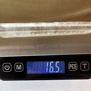 May include: A clear crystal, elongated in shape, rests on a digital scale. The scale shows a weight of 16.5 grams. The scale has a silver surface and a black base with control buttons.