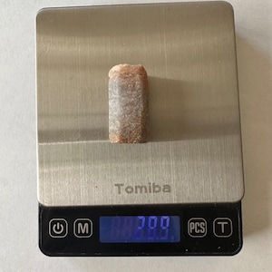 May include: A silver digital scale with a black display showing a weight of 0.200 grams. The scale has a power button, a mode button, a "PCS" button, and a "T" button. A small, brown, rectangular rock is sitting on the scale.