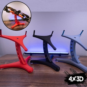Könnte beinhalten: Drei 3D-gedruckte Drohnenständer in Rot, Schwarz und Blau. Die Ständer haben eine gebogene Basis und eine dreizackige Oberseite. Die Ständer sind so konzipiert, dass sie eine Drohne an Ort und Stelle halten.