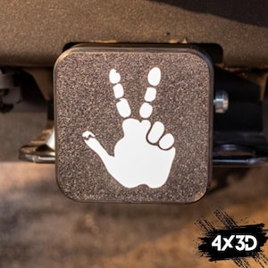 Puede incluir: Una tapa de enganche cuadrada negra con una mano blanca haciendo el signo de la paz. El texto "4x3D" es visible en la esquina inferior derecha de la imagen.