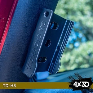 May include: A black plastic radio mount with the text "TLD RADIO H8" printed on it. The mount is attached to a vehicle seat.