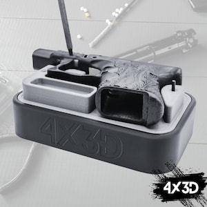 May include: A black plastic 3D printed gun frame sits in a grey and black 3D printed mould. The mould has the text "4X3D" printed on the side.