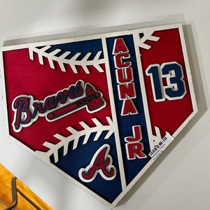 Puede incluir: Un plato de home en madera con un diseño de béisbol rojo y azul. El plato presenta el logotipo de los Atlanta Braves, el nombre "Acuna Jr." y el número "13".