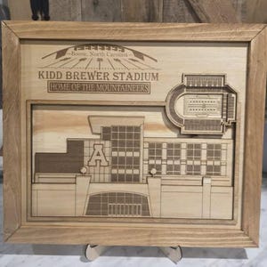 May include: Wooden framed artwork featuring a detailed laser-cut depiction of Kidd Brewer Stadium, Boone, North Carolina. The artwork includes the stadium, buildings, and the text "Kidd Brewer Stadium Home of the Mountaineers". The frame is made of light-colored wood.