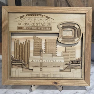 May include: Wooden framed artwork featuring a laser-cut depiction of Acrisure Stadium, home of the Steelers, in Pittsburgh, Pennsylvania. The artwork includes the stadium, city skyline, and the stadium's name.