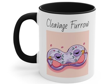 Puede incluir: Taza de cerámica blanca con asa y borde negros. La taza tiene un fondo rosa con una ilustración de dibujos animados de una célula con un sujetador rosa. El texto "Cleavage Furrow" está impreso en negro en la parte superior de la taza.