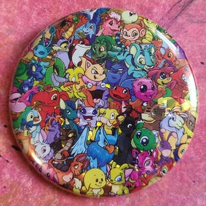 Op de afbeelding: Een ronde knoop met een kleurrijk ontwerp van cartoonfiguren. De knoop heeft een glanzende afwerking en toont een veelheid aan wezens in verschillende kleuren. De achtergrond is een roze, getextureerd oppervlak.