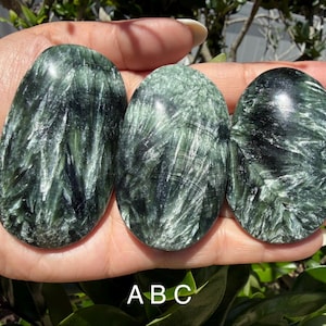 May include: Three polished seraphinite stones in varying sizes, held in a hand. The stones are oval-shaped with a dark green and silver feathery pattern. The stones are labeled A, B, and C.