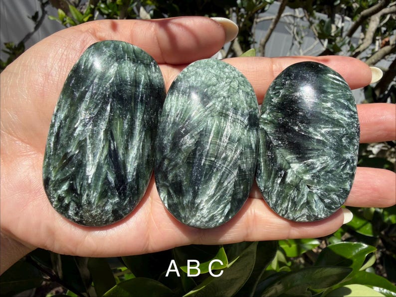 May include: Three polished seraphinite stones in varying sizes, held in a hand. The stones are oval-shaped with a dark green and silver feathery pattern. The stones are labeled A, B, and C.
