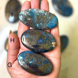 May include: A collection of polished oval-shaped stones with a mix of blue, teal, and gold colors. The stones have a smooth, glossy finish and are held in a hand. The stones are likely used for decorative or metaphysical purposes.