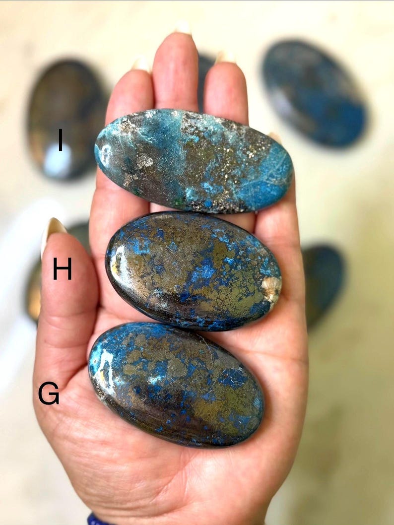 May include: A collection of polished oval-shaped stones with a mix of blue, teal, and gold colors. The stones have a smooth, glossy finish and are held in a hand. The stones are likely used for decorative or metaphysical purposes.