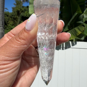 May include: A clear, double-terminated quartz crystal point with a faceted, hexagonal shape. The interior of the stone features visible internal fractures and inclusions, with a small, distinct rainbow-colored light refraction near the center.