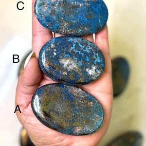 May include: Three oval-shaped stones with a blue and gold color scheme are held in a hand. The stones have a polished, smooth surface. A blue beaded bracelet is visible on the wrist.
