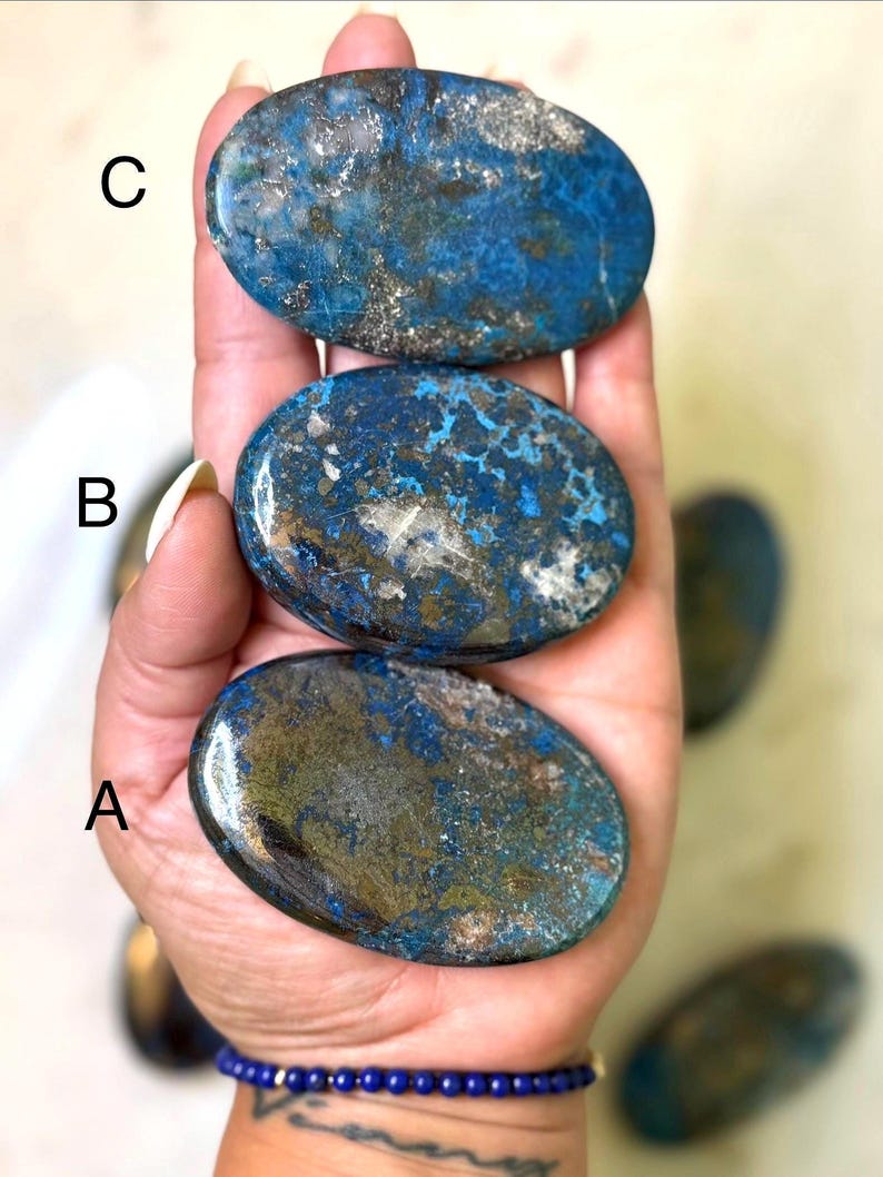 May include: Three oval-shaped stones with a blue and gold color scheme are held in a hand. The stones have a polished, smooth surface. A blue beaded bracelet is visible on the wrist.